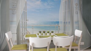 Un área de comedor frente a la playa, con sillas y mesas blancas, vista al mar y un velero a lo lejos.