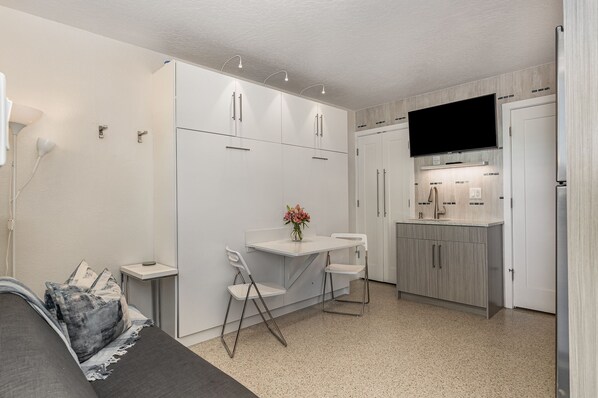 A compact, modern kitchen with white cabinetry, a built-in TV, and a small dining area with a foldable table and chairs.