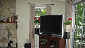 Uma sala de estar com lareira de pedra, uma grande TV de tela plana sobre um suporte de madeira, e vista para o verde através de uma janela ampla.