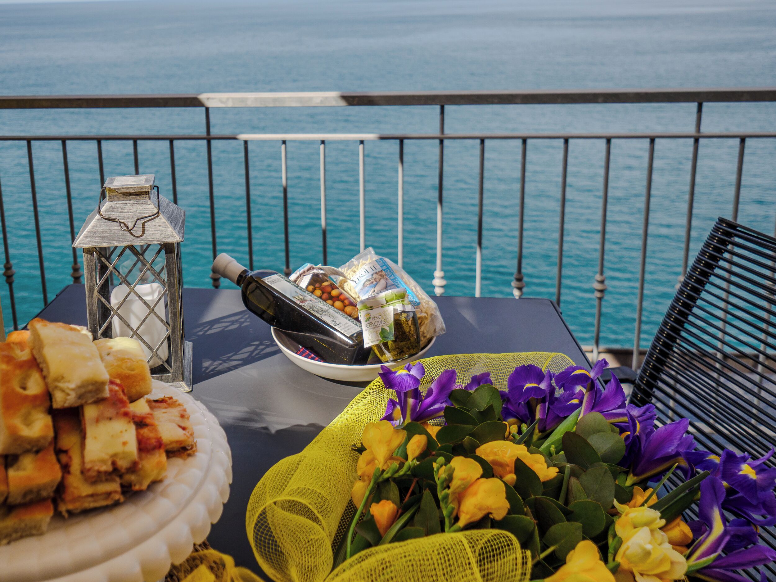 Une table dressée avec de la nourriture, des fleurs et une bouteille, sur un balcon offrant une vue sur la mer.