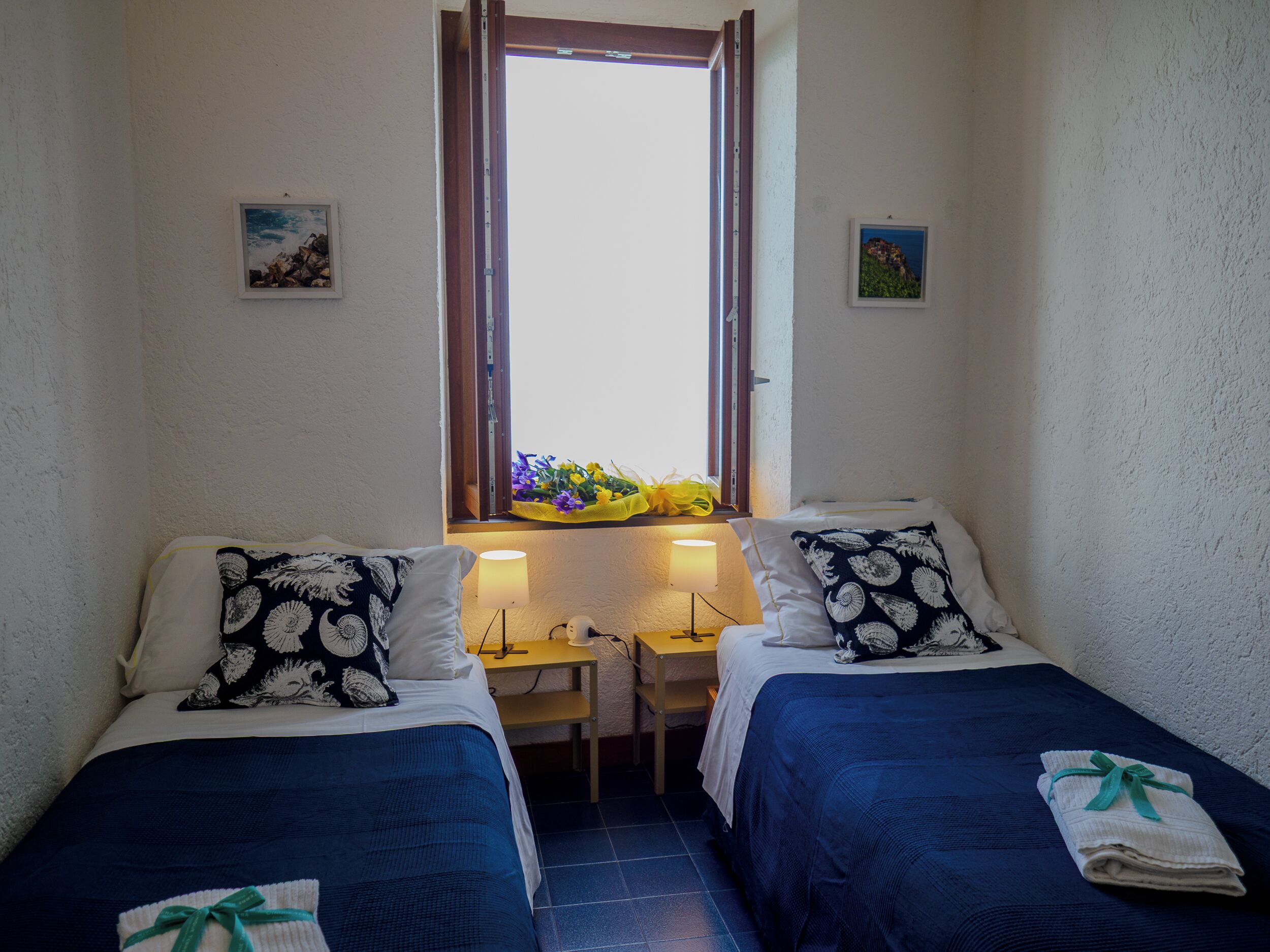 A small, cozy bedroom with two beds, each with a blue bedspread and a pillow with a black and white pattern. There is a window with a view of the sky and a small table with a lamp between the beds.