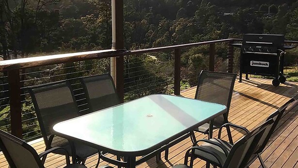 A deck with a glass-top table and chairs, overlooking a forested hillside.