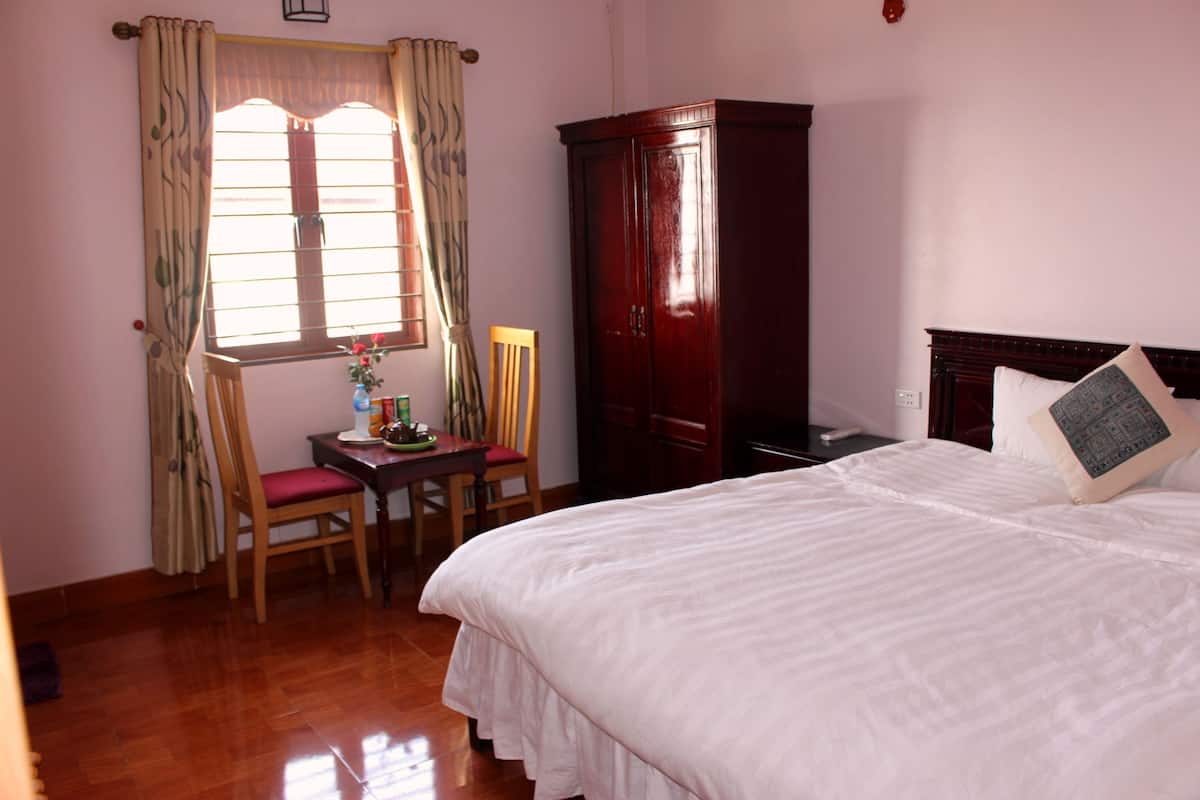 A bedroom with a bed, a wooden wardrobe, a desk with a chair, and a window with curtains.