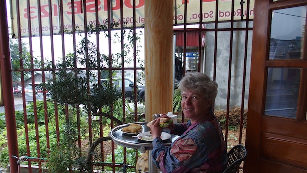 Een vrouw zit aan een tafel met eten en een drankje, buiten een gebouw met een bord in een vreemde taal.
