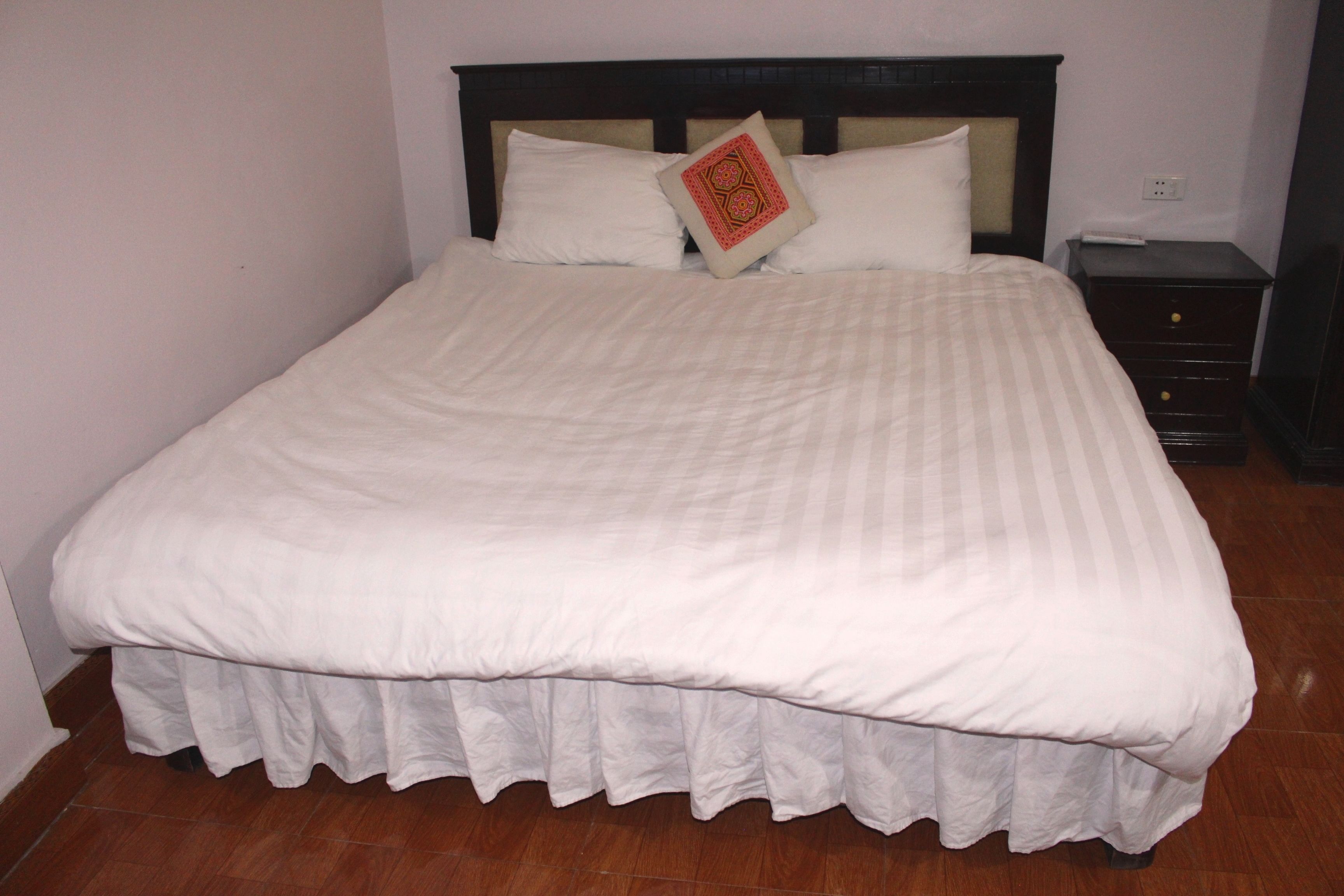 A neatly made bed with a white bedspread and a decorative pillow, a wooden nightstand with a drawer, and a wall socket.