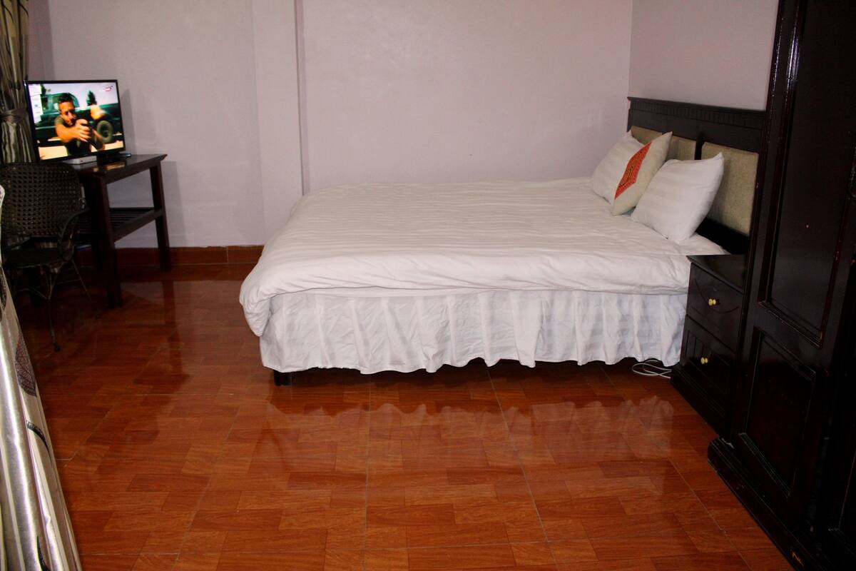 A neatly made bed with a white bedspread, a wooden nightstand with a television, and a wooden floor.