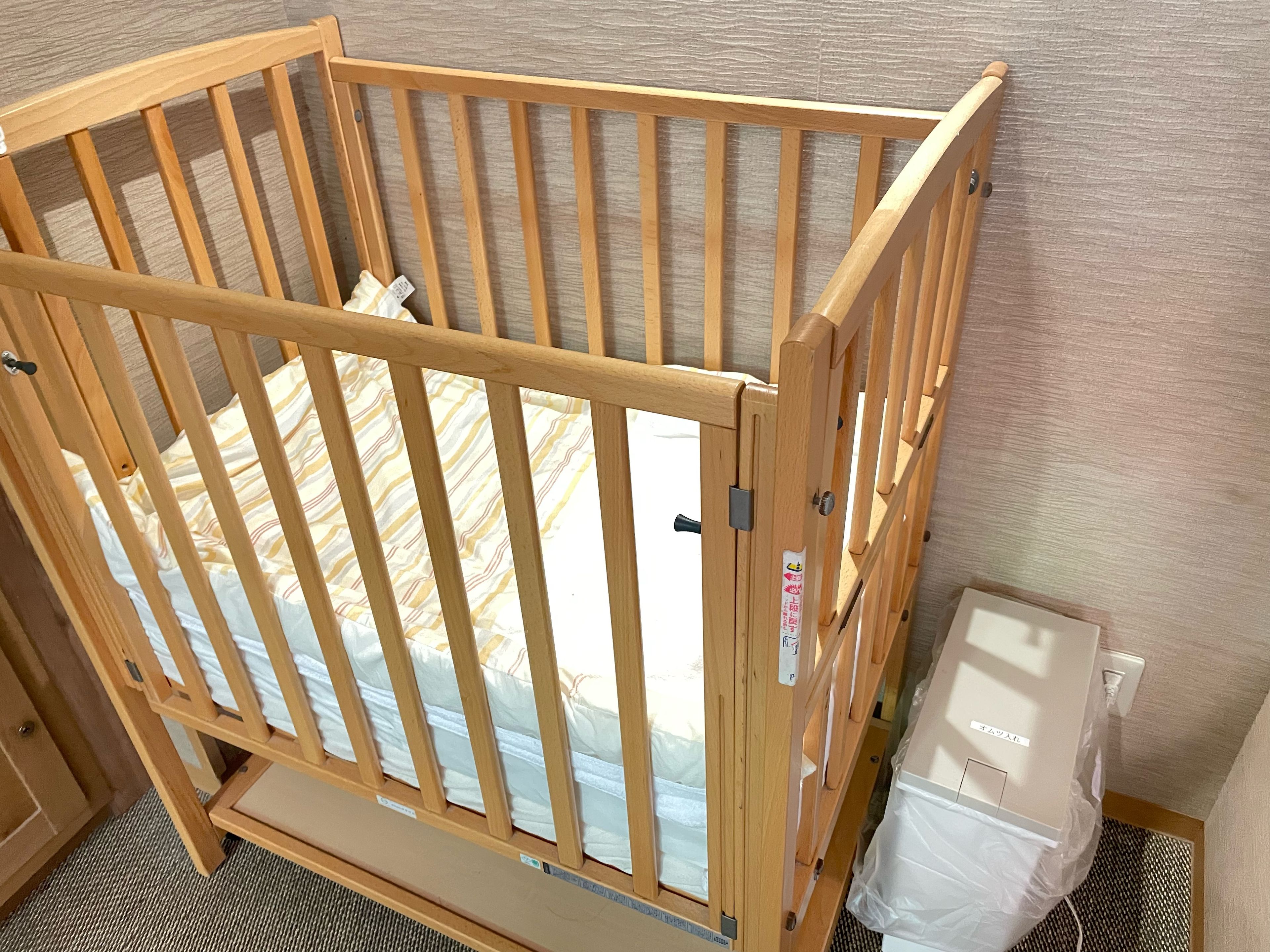 A wooden crib with a mattress, a wall-mounted outlet, and a plastic bag on the floor.