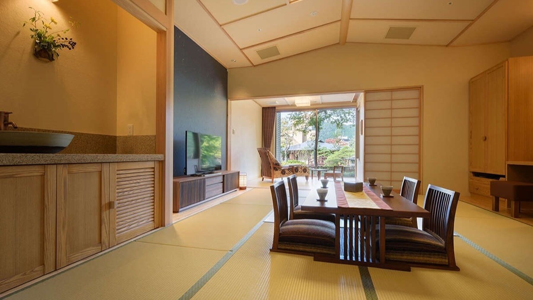 Una habitación tradicional japonesa con suelo de tatami, una mesa baja con cojines, un televisor y puertas correderas que dan a un jardín.