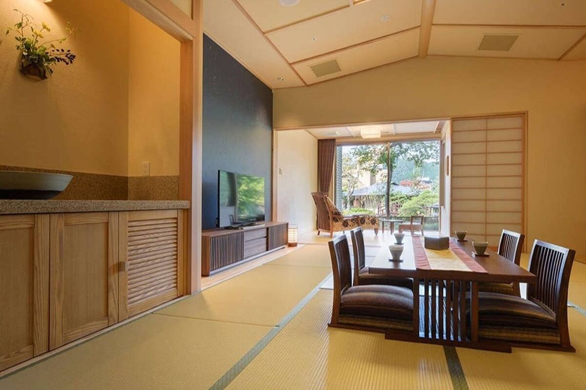 A traditional Japanese-style room with tatami flooring, a low table with cushions, a television, and sliding doors leading to a garden.