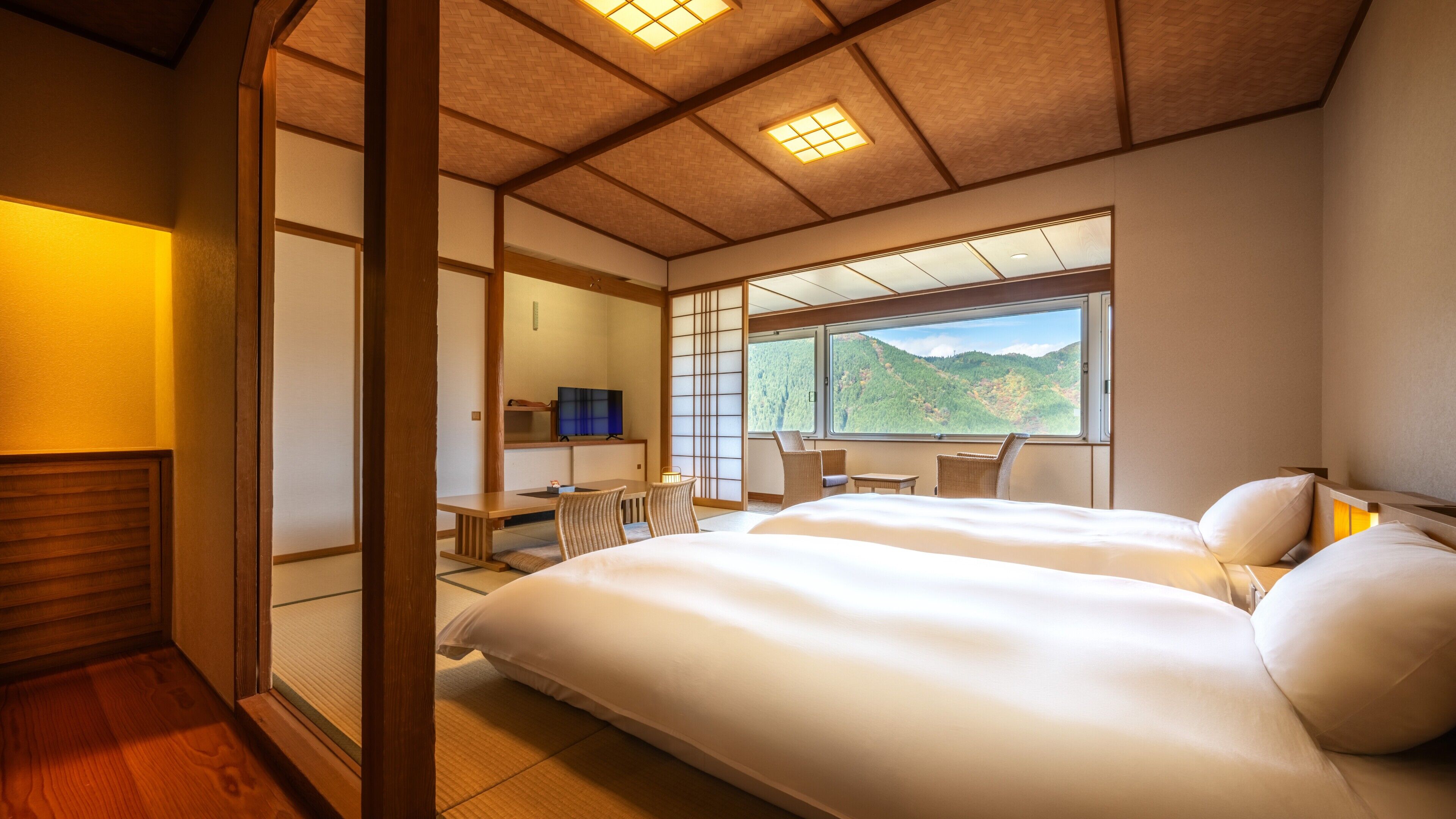 Habitación de hotel japonesa tradicional con suelo de tatami, una cama grande, un escritorio con silla y vistas a un paisaje montañoso.