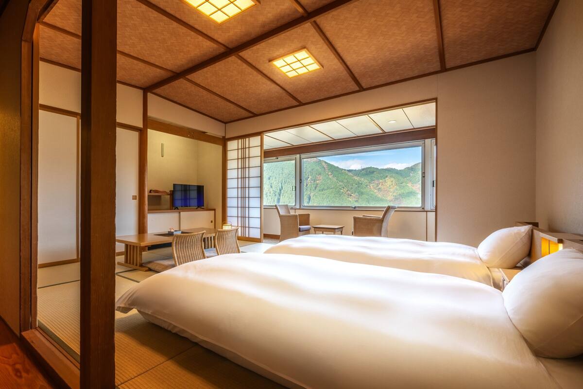 A traditional Japanese-style hotel room with tatami flooring, a large bed, a desk with a chair, and a view of a mountainous landscape.
