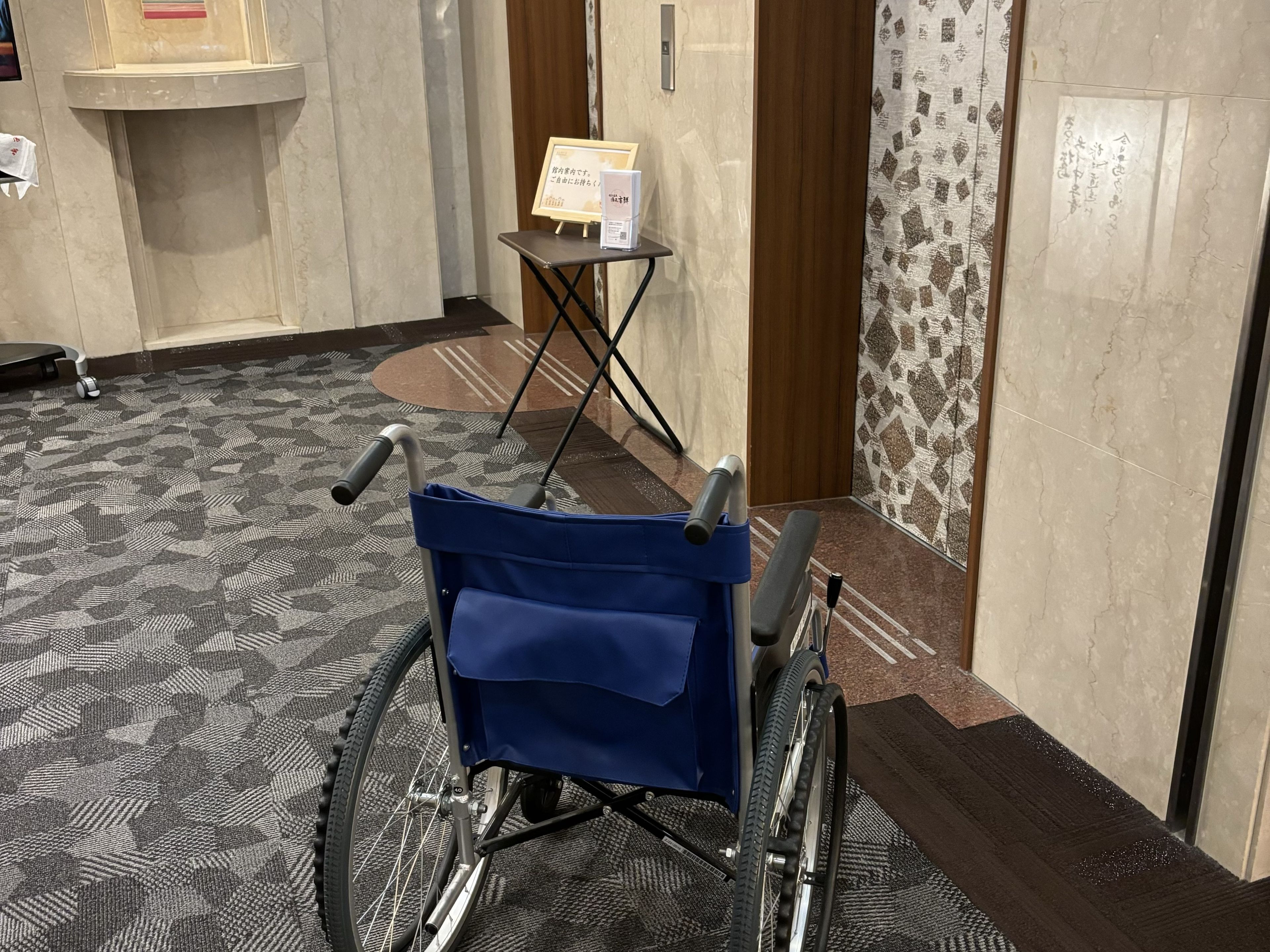 A wheelchair in a hotel hallway with a patterned carpet, a small table with a sign, and a door with a patterned wall.