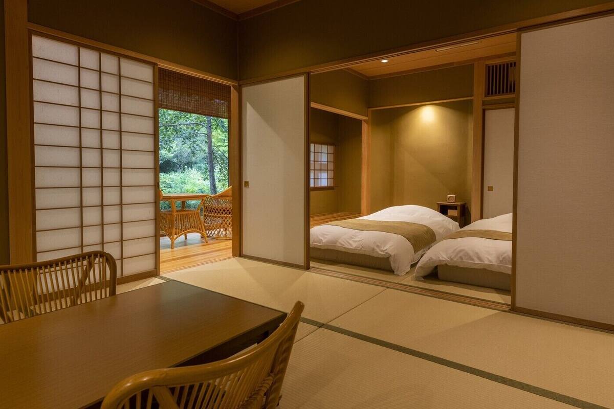 A traditional Japanese room with tatami flooring, a low table with chairs, and a sliding door leading to a bed area.