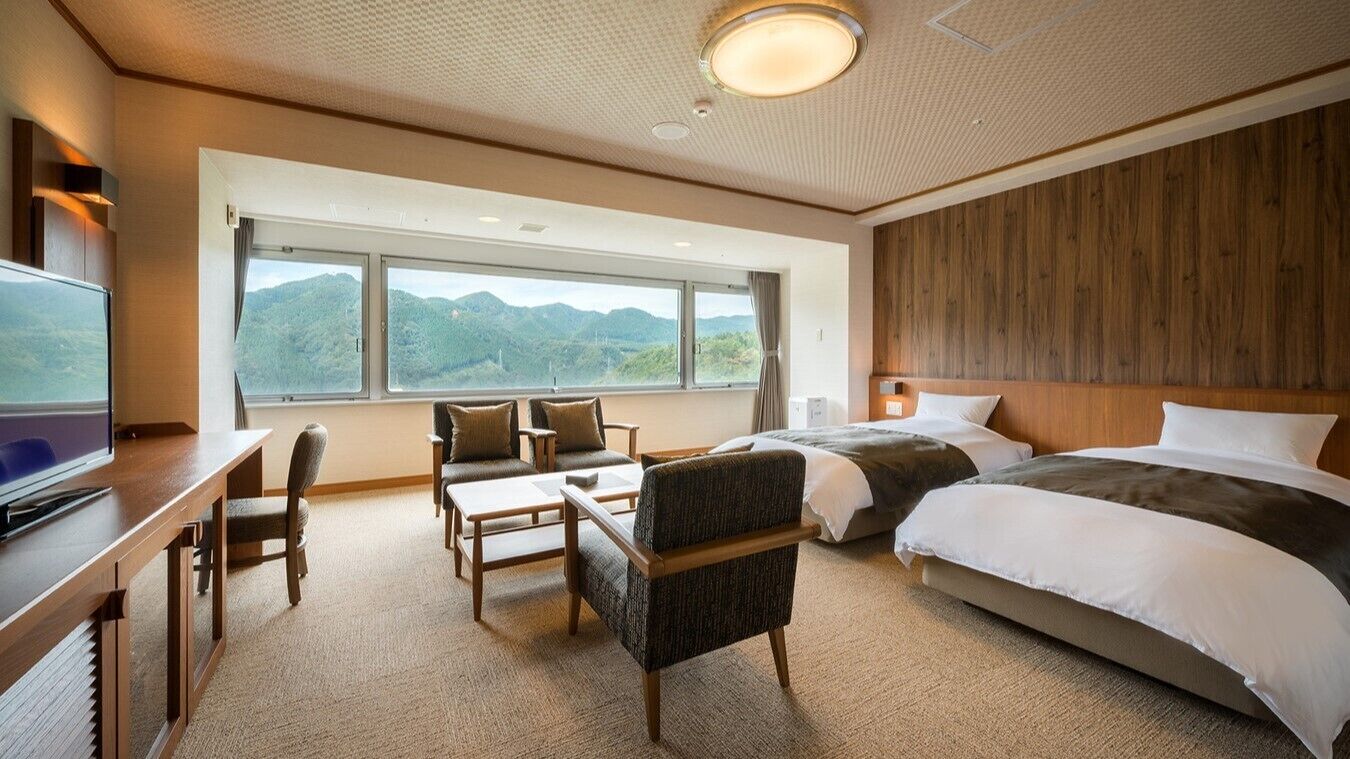Habitación de hotel con un ventanal grande, un escritorio con televisor, dos camas y una zona de estar con sillas y una mesa.