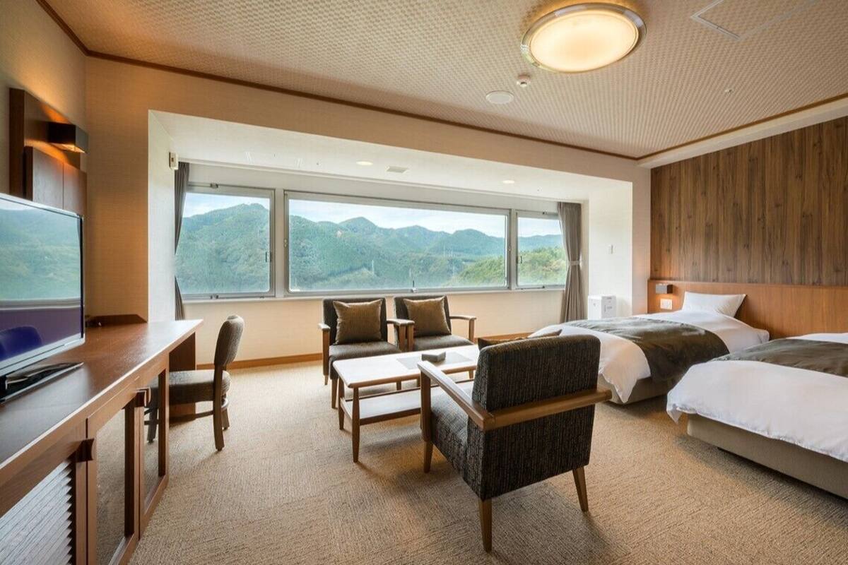 A hotel room with a large window, a desk with a TV, two beds, and a seating area with chairs and a table.