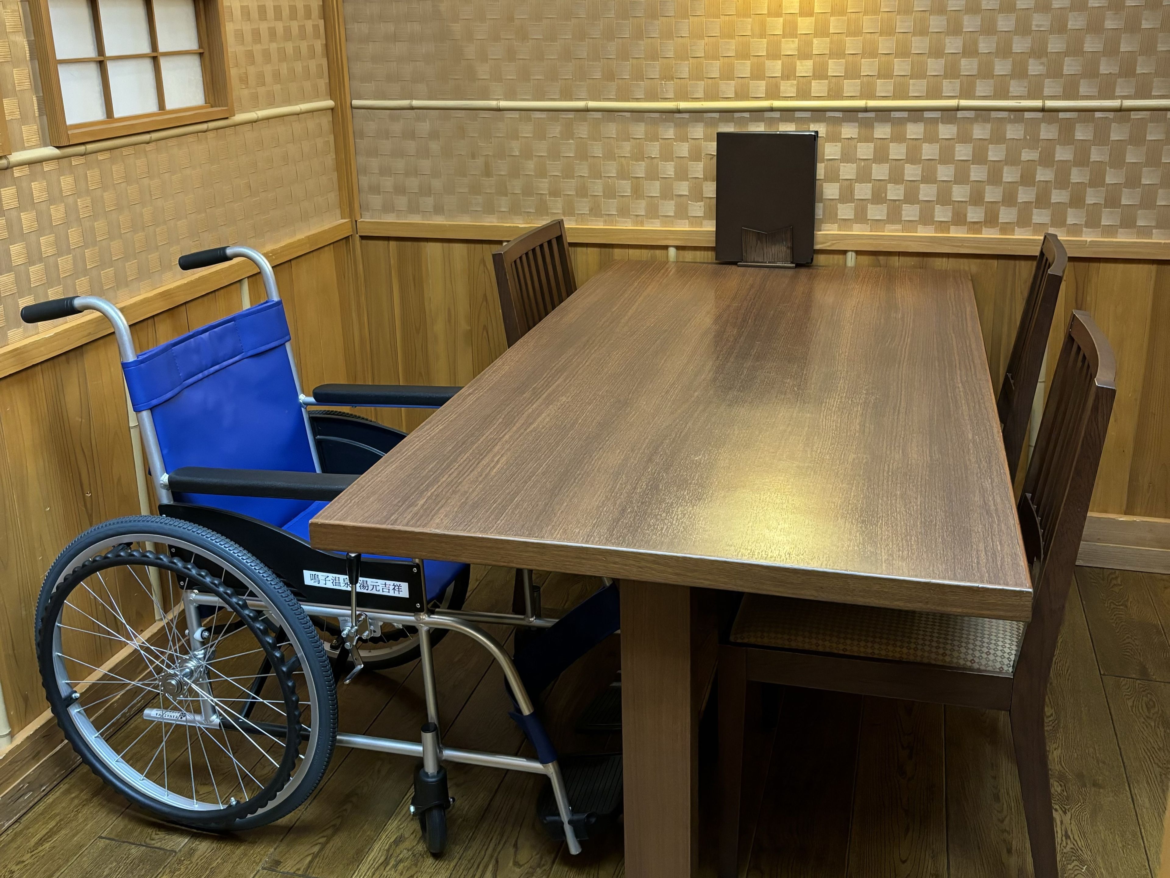 Mesa accesible en silla de ruedas con acabado de madera, ubicada en una habitación con paneles de madera y pared de bambú trenzado.