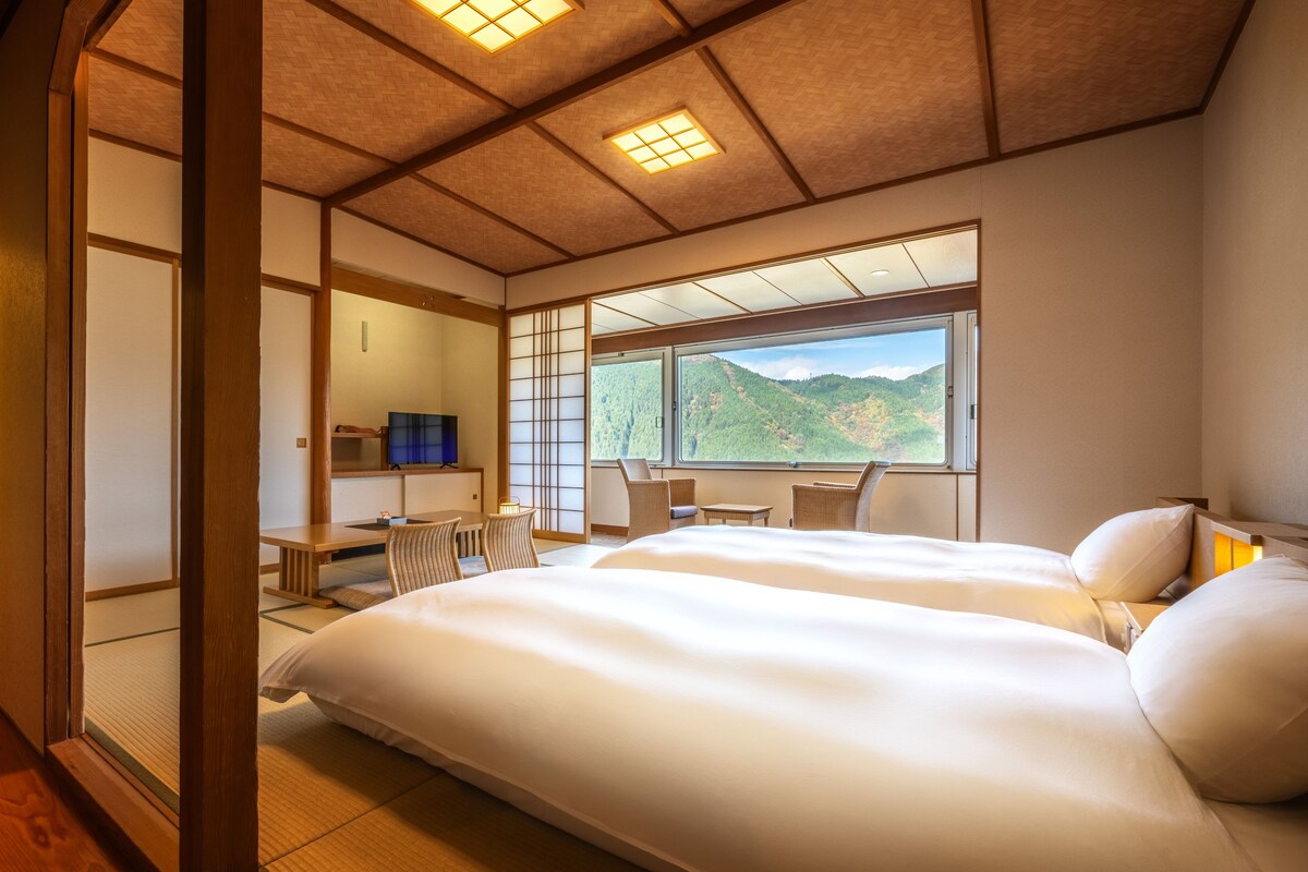 A traditional Japanese-style hotel room with tatami flooring, a large bed, a desk with a chair, and a view of a mountainous landscape.