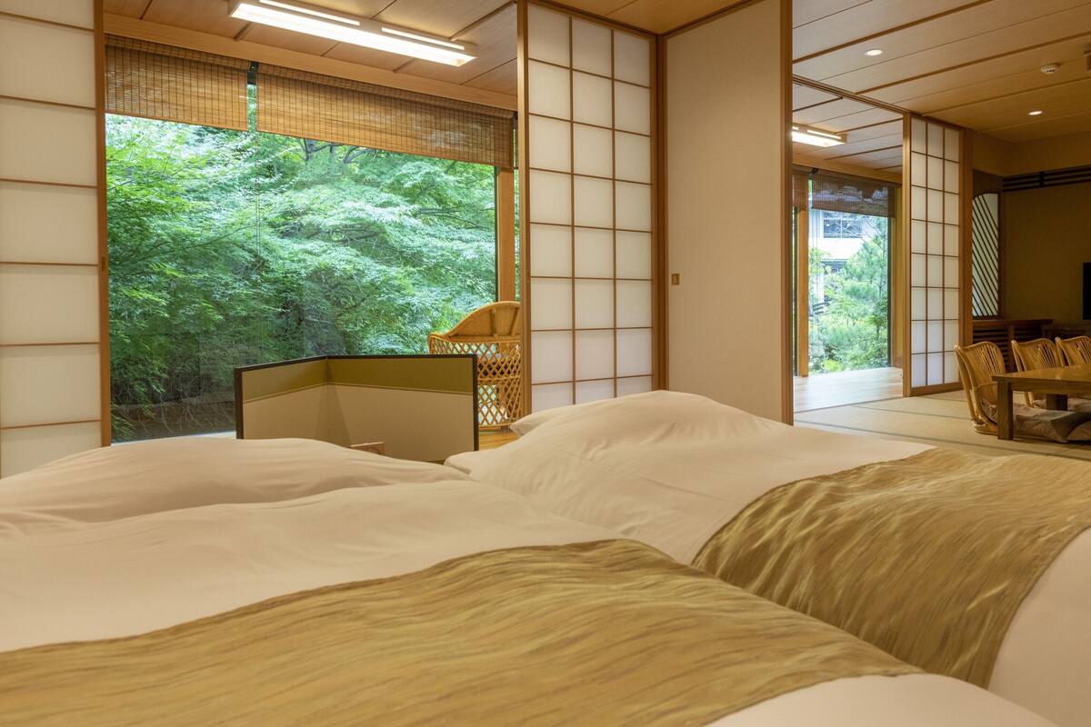 A traditional Japanese-style bedroom with tatami flooring, a bed with white bedding, a wooden desk, and a view of a lush green forest.