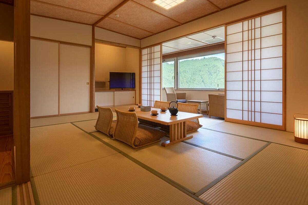 A traditional Japanese room with tatami flooring, a low table with cushions, a television, and a sliding door with a view of greenery.