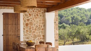 A wooden dining table with chairs and a basket of fruit, set against a stone wall with a view of trees. - Villa Dalt S'Era - Free week from July 13 to 20 (Santa Eulària des Riu)