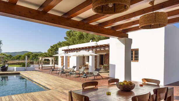 An outdoor pool area with wooden deck and lounge chairs, a dining table with chairs, and a building with white walls and wooden beams in the background. - Villa Dalt S'Era - Free week from July 13 to 20 (Santa Eulària des Riu)