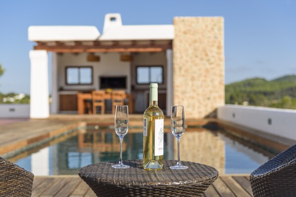 A bottle of wine and two glasses on a wicker table by a poolside, with a building in the background.