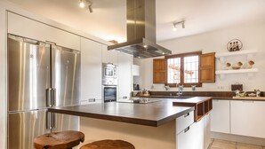 A modern kitchen with stainless steel appliances, a central island with stools, wooden cabinets, and a window with a view. - Villa Dalt S'Era - Free week from July 13 to 20 (Santa Eulària des Riu)