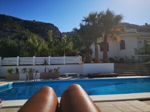 Área de piscina com vista para uma montanha, palmeiras e um muro branco.