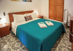 A neatly made bed with a teal bedspread and white pillows, a wooden nightstand with a lamp, and patterned floor tiles.