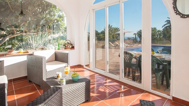 A sunlit patio with wicker furniture, a dining area, and a view of a pool and palm trees. - Villa Garlanda - PlusHolidays (Benissa)