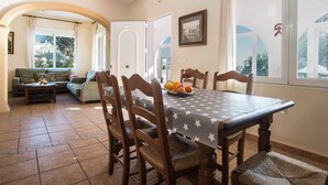 A dining area with a table set for six, surrounded by wooden chairs with woven seats. A bowl of fruit is on the table, and the room has large windows with curtains. - Villa Garlanda - PlusHolidays (Benissa)