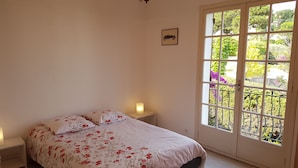 A bedroom with a bed, two bedside lamps, a chandelier, a window with a view of greenery, and a framed picture on the wall.