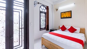 A bedroom with a wooden bed, white bedding, red pillows, a nightstand, a wall-mounted fan, a window with a curtain, and a framed picture on the wall. - Bong House Homestay Hoi An (Da Nang)