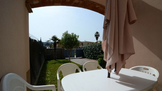 Un patio con una mesa y sillas blancas, una sombrilla y vistas a un jardín y edificios al fondo.