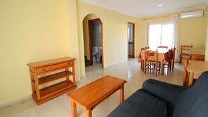 Una sala de estar con un sofá, una mesa de centro de madera, una mesa de comedor con sillas y una zona de cocina visible a través de un arco.