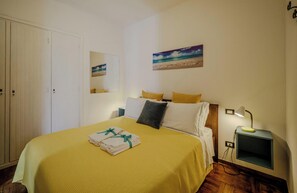 A hotel room with a bed, a nightstand, a lamp, a painting of a beach, and a wardrobe.
