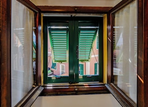 A window with a wooden frame and green shutters, overlooking a street with buildings.