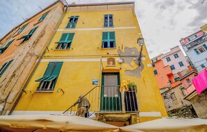 A yellow building with green shutters, a visible door, and a balcony with a railing.
