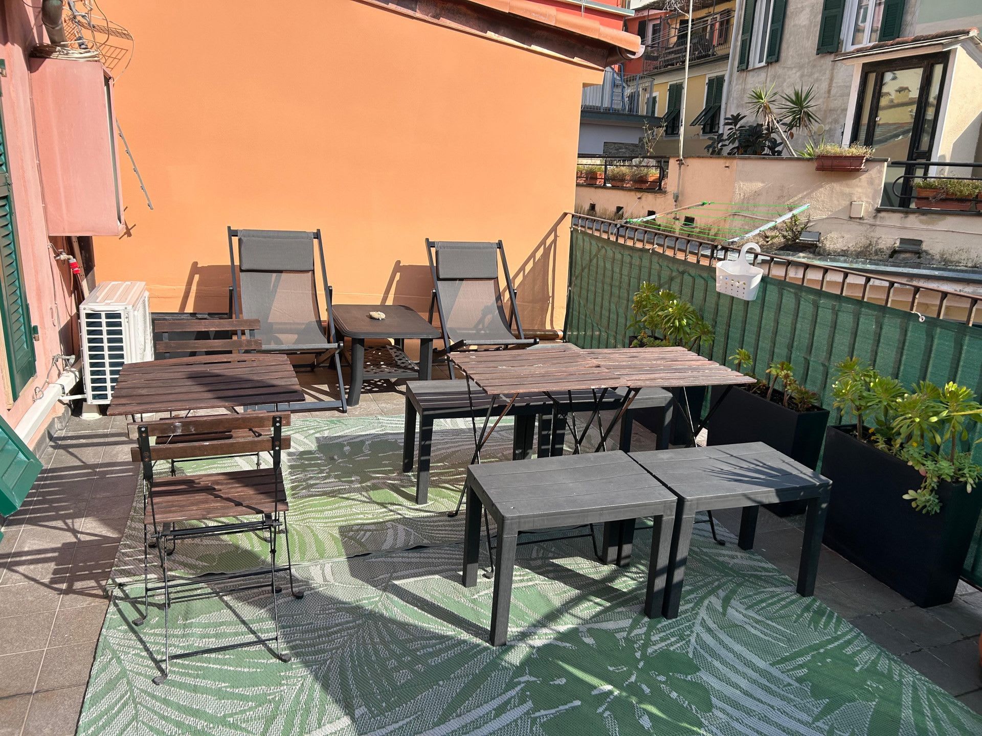 An outdoor patio with wooden tables and chairs, a green patterned rug, and potted plants.