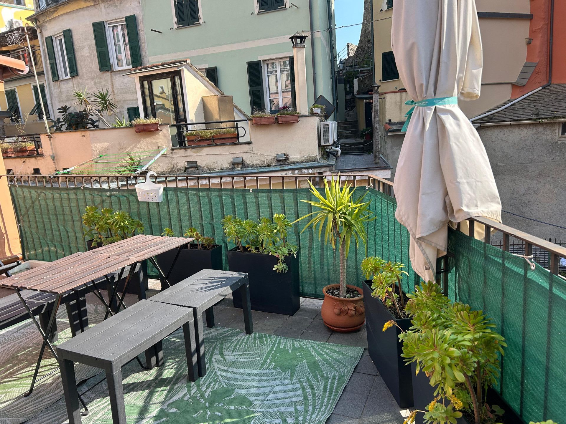 Une terrasse extérieure avec une table et des bancs en bois, entourée de plantes en pot et d’une clôture en treillis vert.
