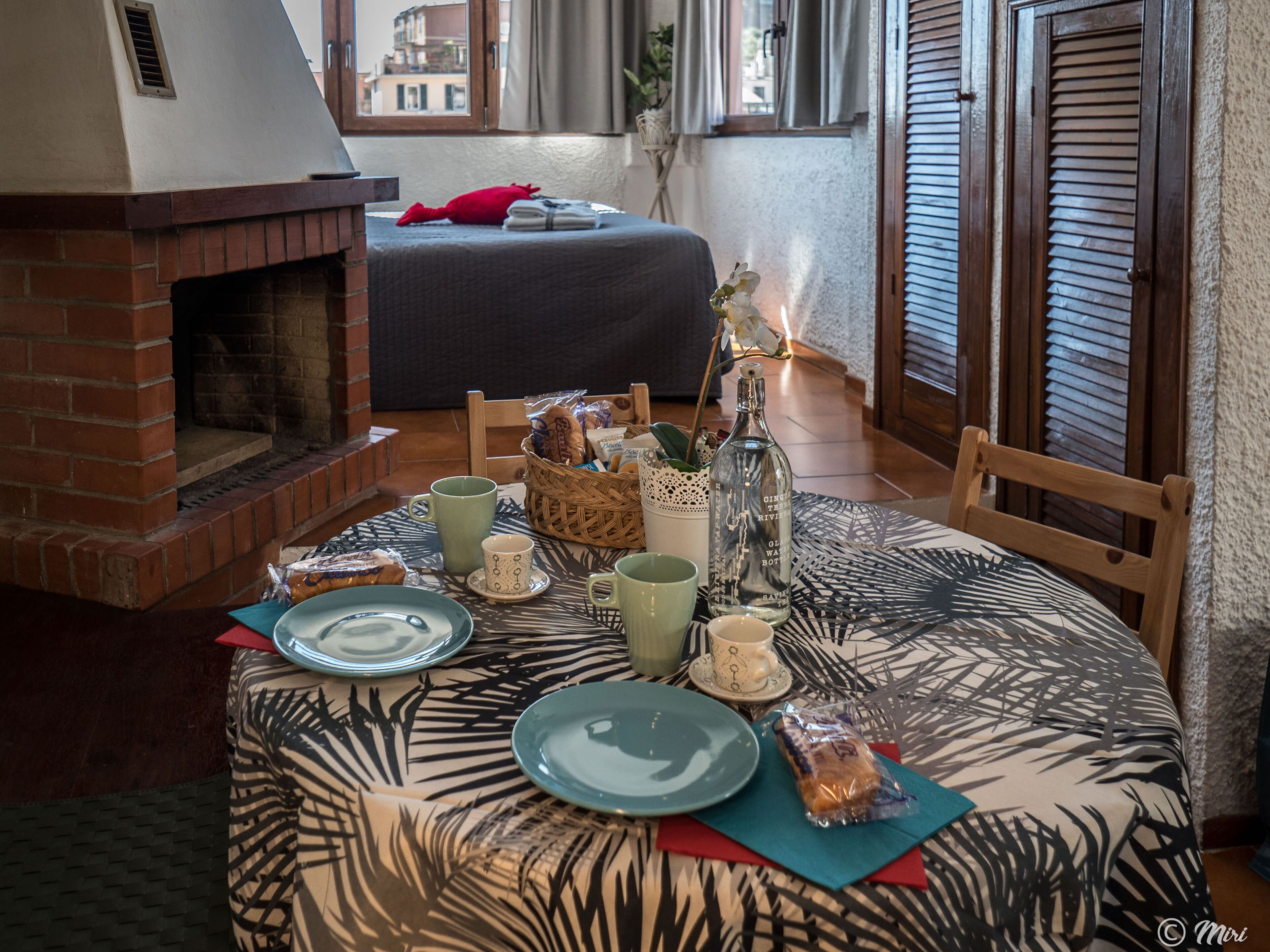 A dining area with a brick fireplace, a table set with plates, cups, and a bottle, and a view of the city through a window.