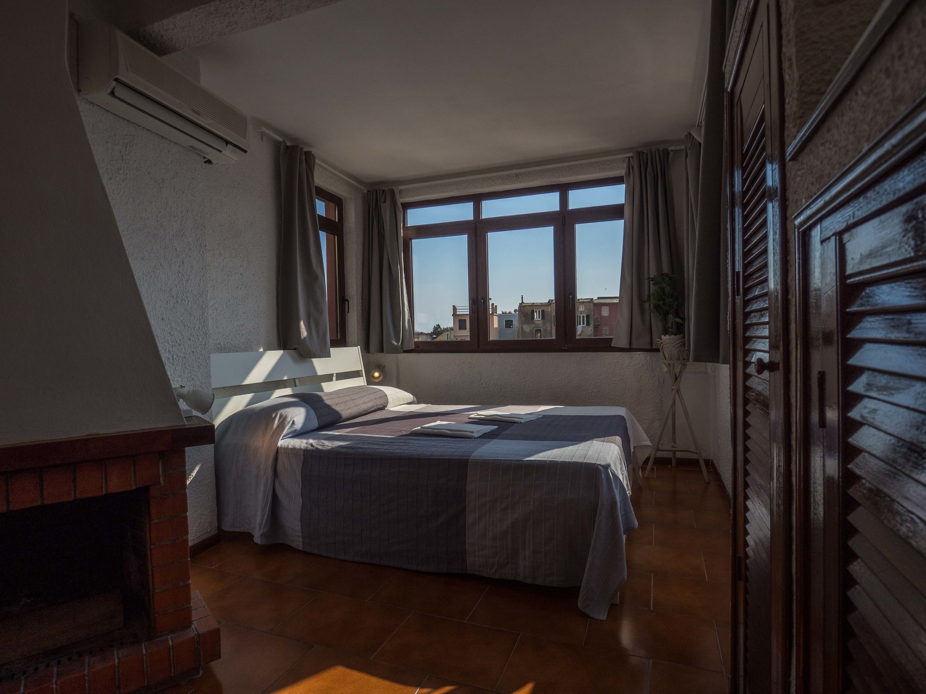 Un dormitorio con cama, chimenea, ventana con cortinas y vista a edificios.