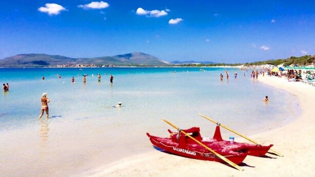 A beach with clear turquoise water, a red boat, and a mountainous backdrop. - ClickSardegna Penthouse Valencia with Sea-View Terraces, Close to the Beaches (Alghero)
