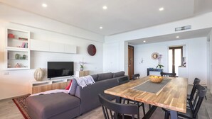 A modern living room with a grey sofa, wooden dining table, and black chairs. A flat-screen TV is mounted on the wall, and there are shelves with decorative items. - ClickSardegna Penthouse Valencia with Sea-View Terraces, Close to the Beaches (Alghero)