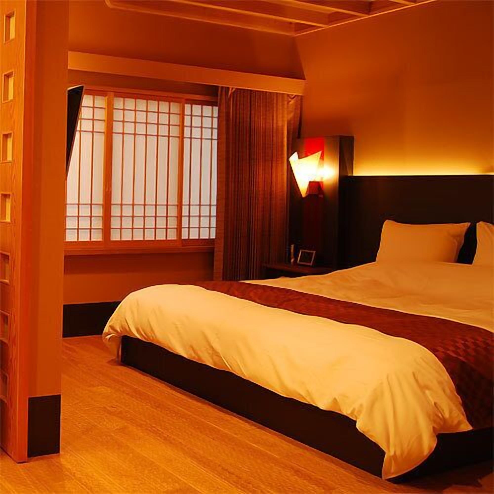 Habitación de hotel con una cama grande, una mesita de noche, una lámpara y una ventana con persianas.