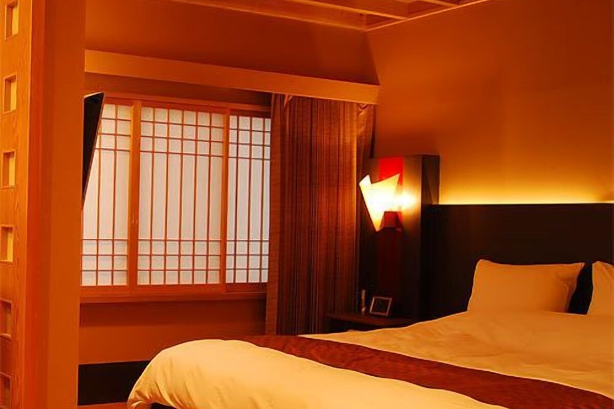 Habitación de hotel con una cama grande, una mesita de noche, una lámpara y una ventana con persianas.