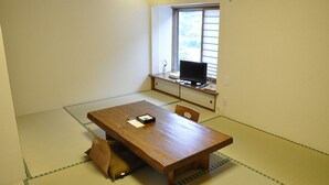Um quarto com piso de tatame, uma mesa de madeira, uma televisão e uma janela com persianas.