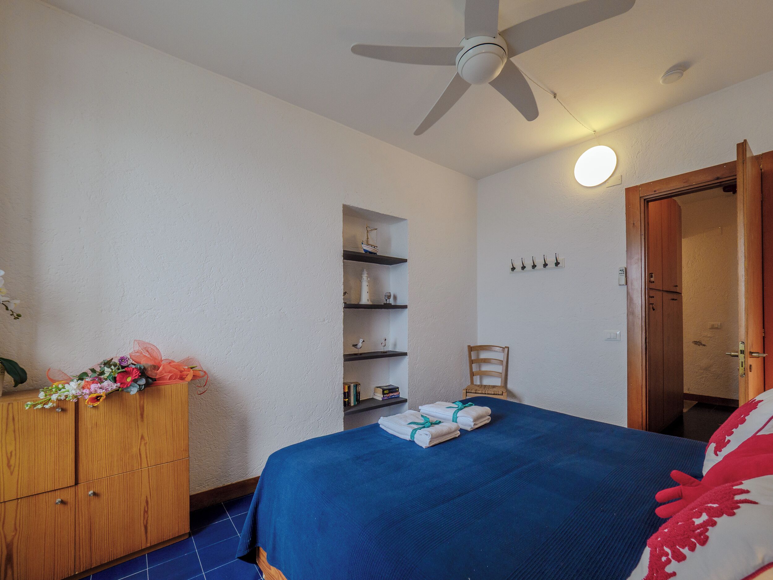 Un dormitorio con cama, cómoda de madera, ventilador de techo, estante en la pared con adornos y una puerta que da a otra habitación.