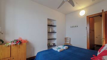 Un dormitorio con cama, cómoda de madera, ventilador de techo, estante en la pared con adornos y una puerta que da a otra habitación.