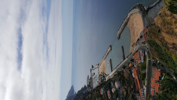 A coastal town with a prominent pier, a beach, and a road leading up to a hill with buildings. - Casa do Leme - Rustic House (Funchal- Madeira)